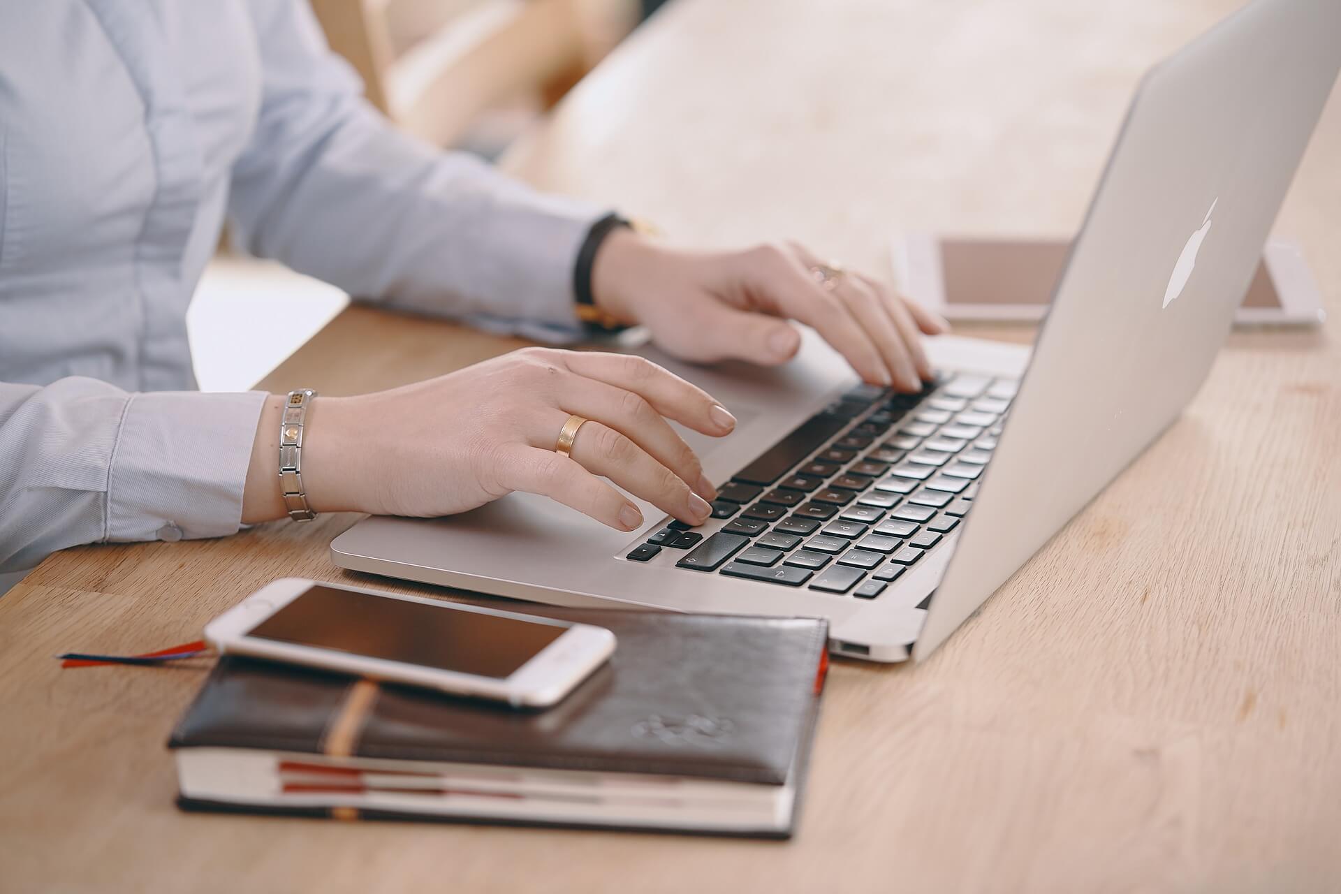 Woman typing on keyboard with phone and notepad next to her laptop.